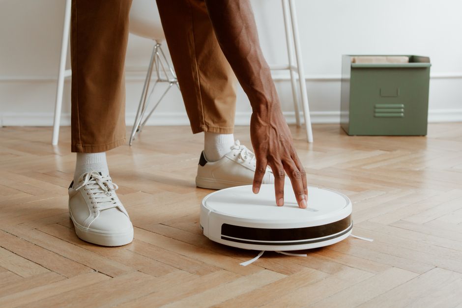 A man's hand adjusting a robot vacuum on a wooden floor in a modern home interior.