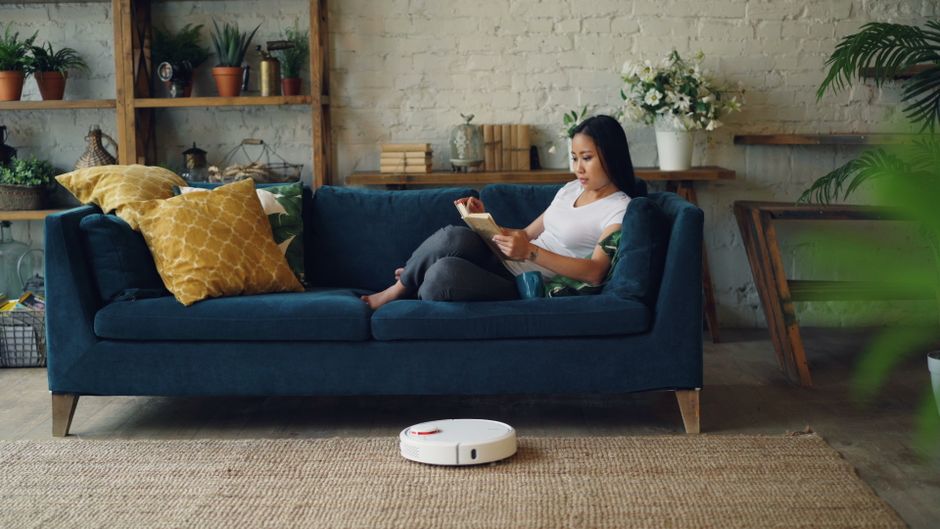 Woman reading on sofa while a robot vacuum cleans the floor in a cozy living room.